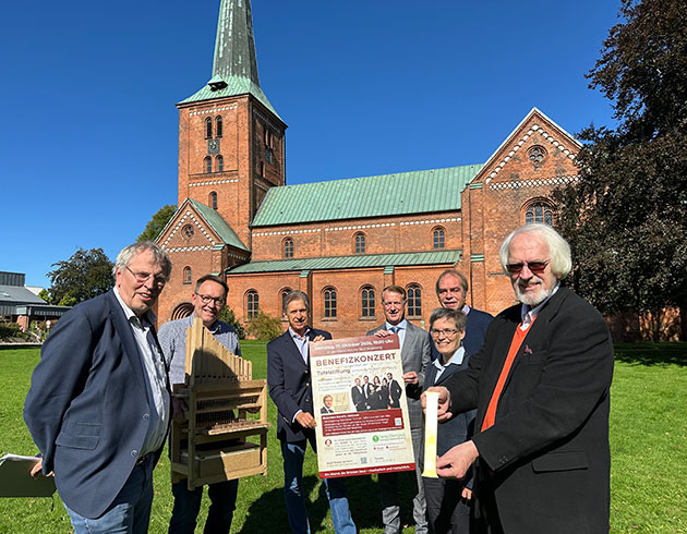 Gruppe vor Kirche mit Plakat für Benefizkonzert.