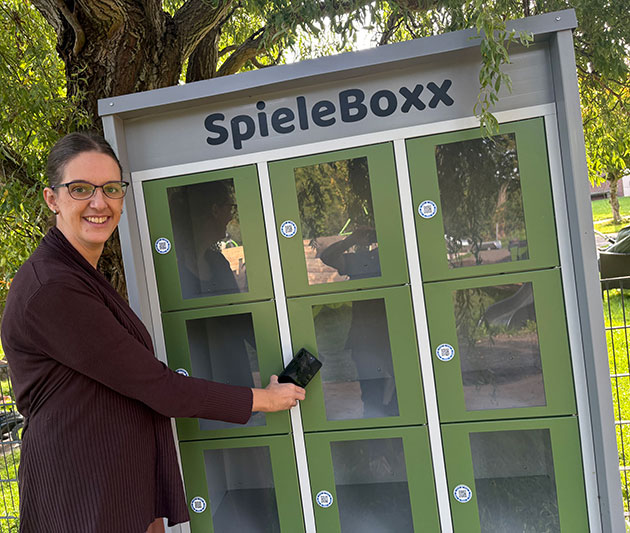 Frau steht lächelnd vor grüner SpieleBoxx mit Handy.