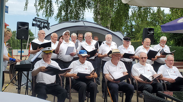 Männerchor singt im Freien, Bäume im Hintergrund.