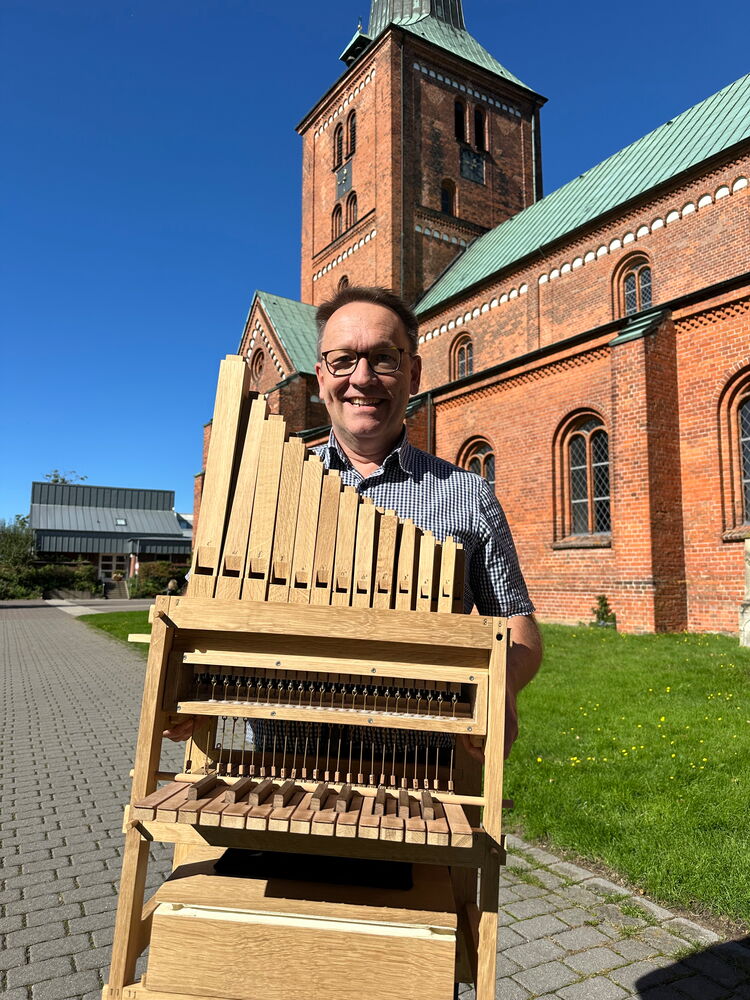 Mann hält ein hölzernes Musikinstrument vor einer Backsteinkirche.