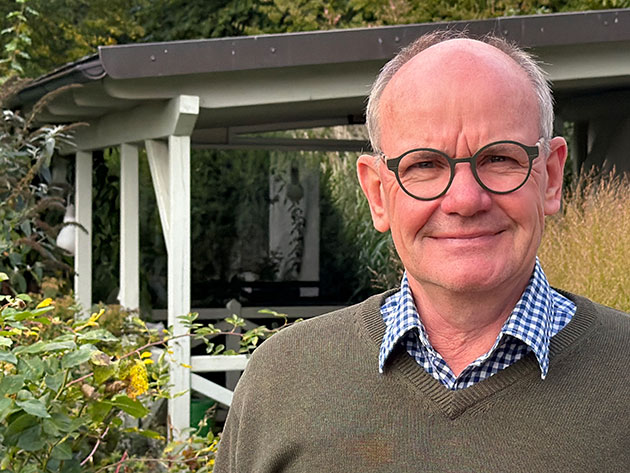 Mann mit Brille vor Gartenhaus und Pflanzen.