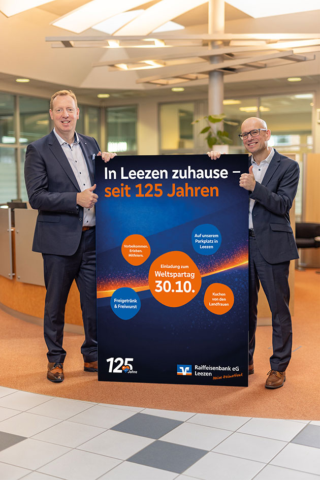 Zwei Männer halten ein Plakat zum Weltspartag in einer Bankfiliale.