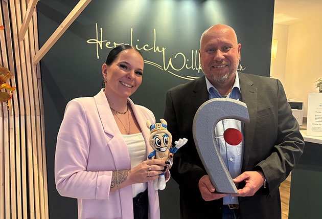 Zwei Personen mit einem Maskottchen und einer Trophäe in der Hand.
