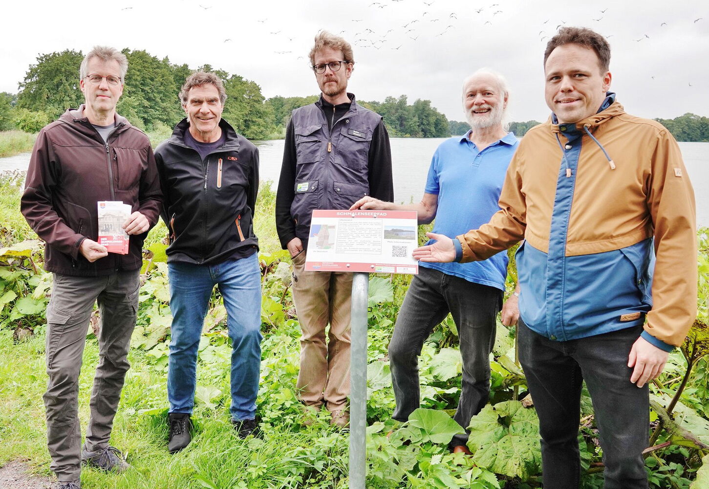 Fünf Männer stehen draußen um ein Schild am See.