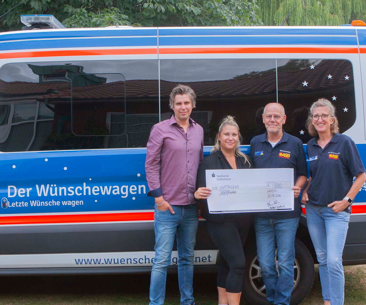 Vier Personen halten einen großen Scheck vor einem Wünschewagen.